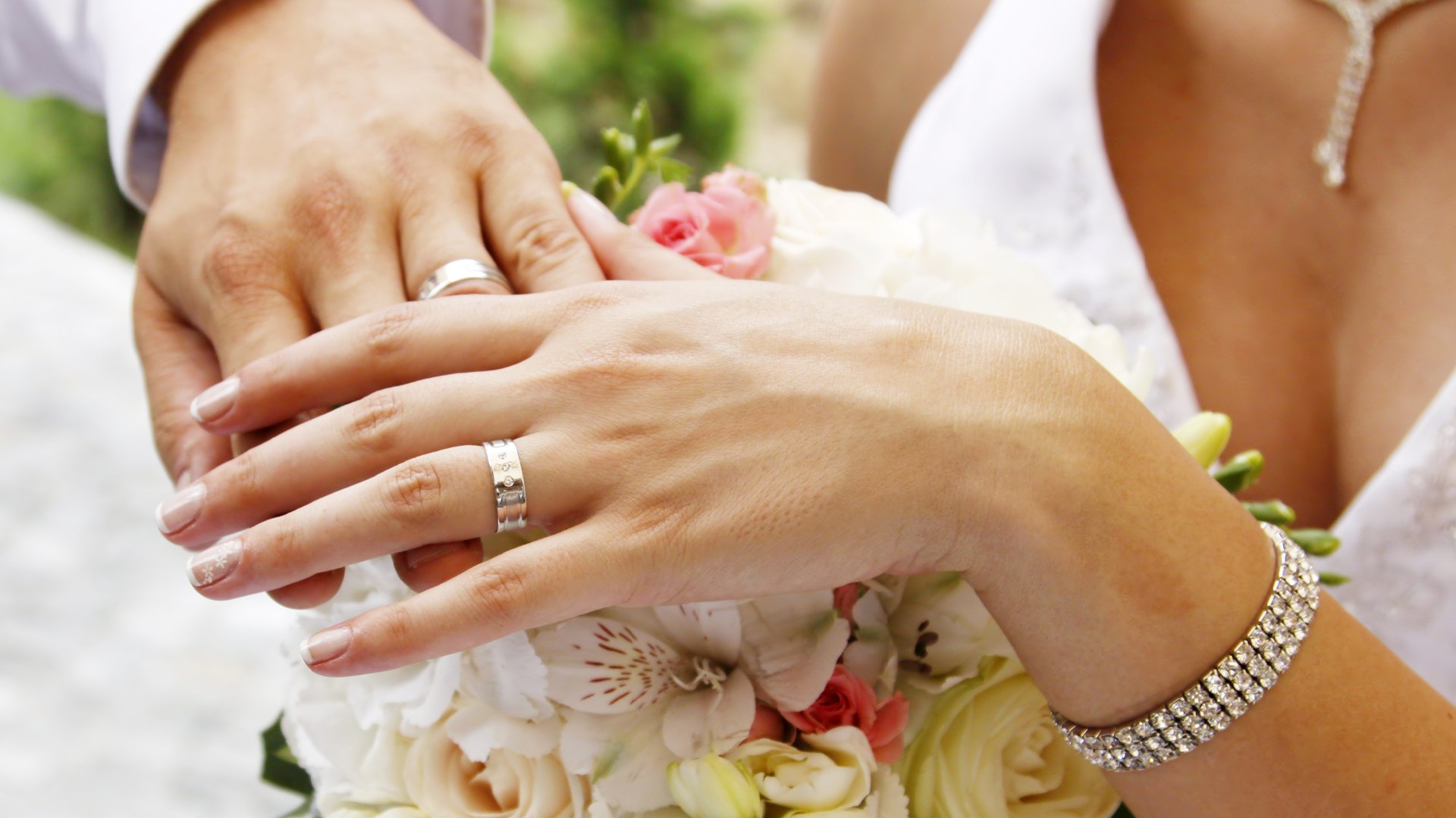 Mani di sposi appoggiate sul bouquet, con le fedi nuziali al dito e un bracciale di brillanti