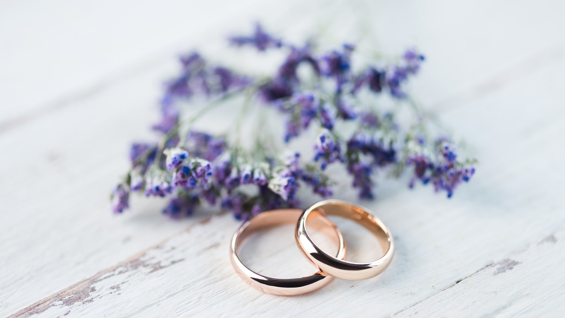 Due fedi nuziali in oro rosa appoggiate su un tavolo di legno bianco accanto a un mazzetto di fiori di lavanda